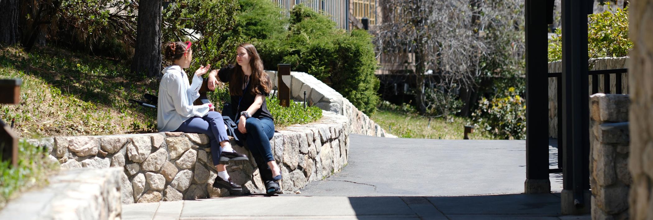 Two young women sit on a short stone wall and engage in conversation.